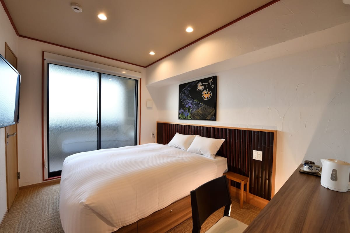 Hotelkamer met een groot bed, een houten hoofdbord, een schilderij aan de muur, een schuifglazen deur en een klein tafeltje met een waterkoker.