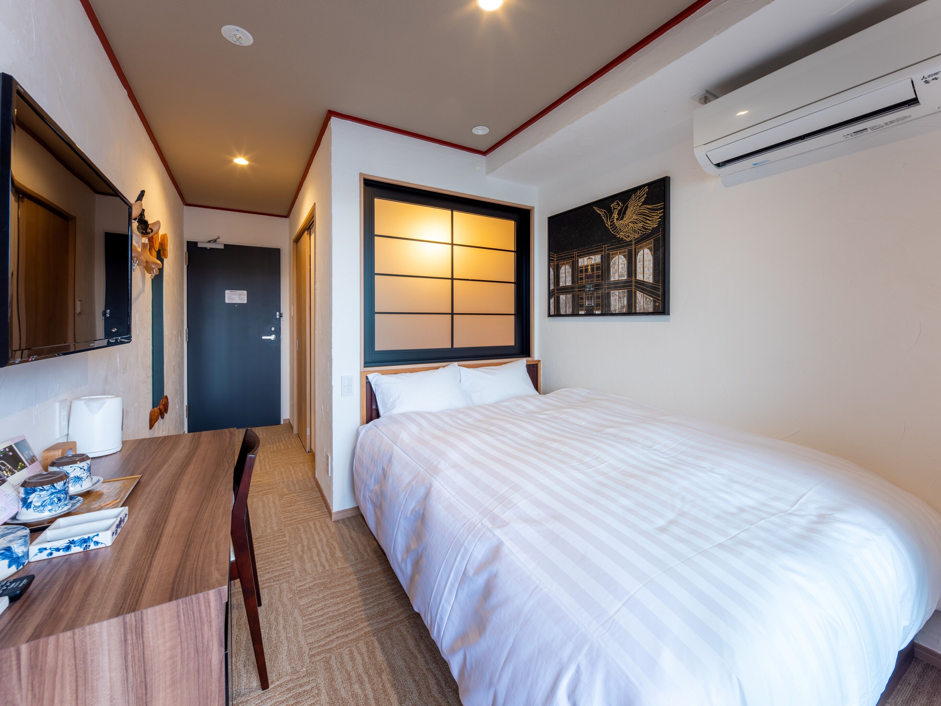 A hotel room with a bed, a desk, a chair, a mirror, an air conditioner, and a door.