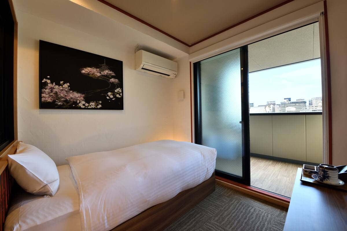 Hotelkamer met een bed, een wandairconditioner, een schuifdeur naar een balkon en een schilderij van kersenbloesems.