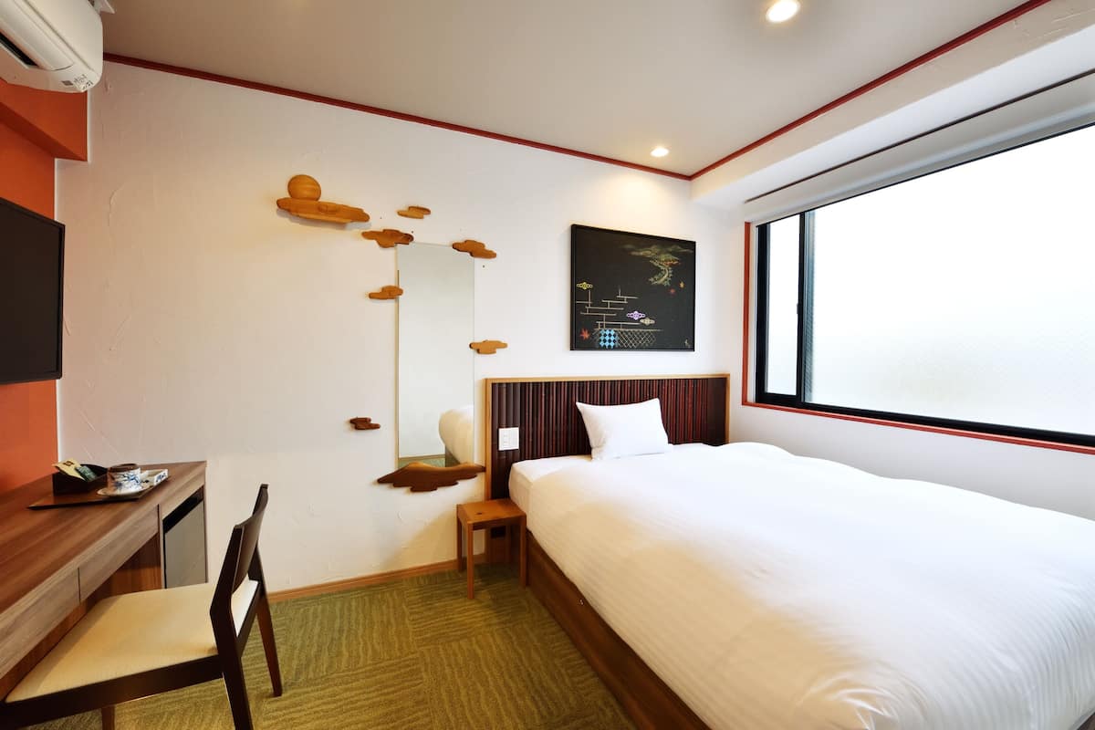 Quarto de hotel com cama, escrivaninha, cadeira, mesinha, espelho fixado na parede e janela.