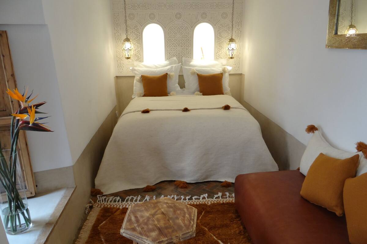 A bedroom with a bed, two wall-mounted lamps, a wooden headboard, a brown sofa, and a decorative mirror.