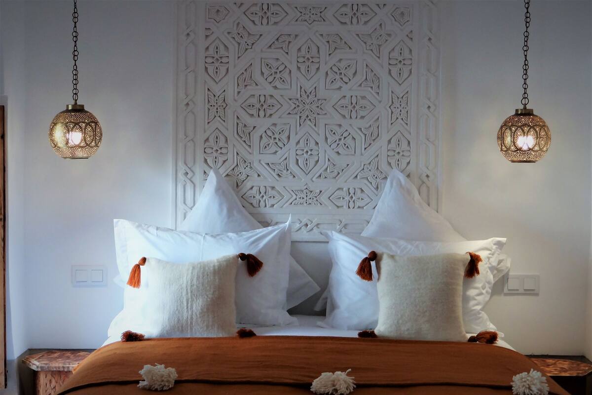 A neatly made bed with white and brown bedding, two hanging lamps, and a decorative wall panel.