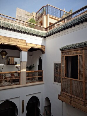 A courtyard with a traditional architectural style, featuring a wooden balcony with intricate lattice work, a tiled roof, and a small wooden window with a lattice design.