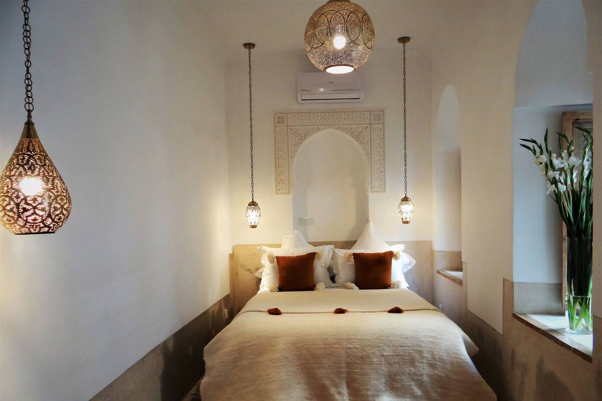 A neatly arranged bedroom with a bed, hanging lamps, a mirror, and a window with a plant.