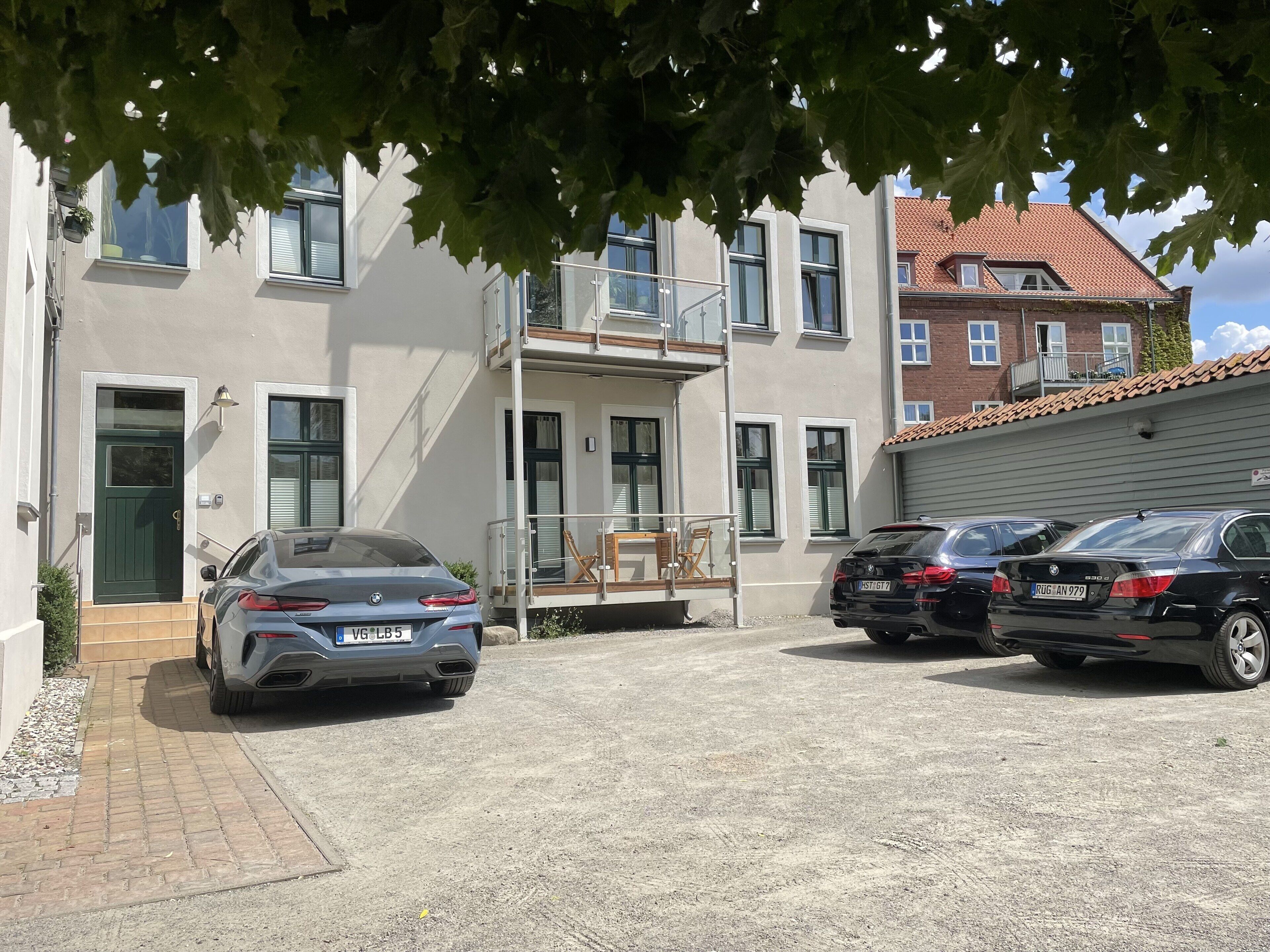 Ein Wohnhaus mit gepflasterter Auffahrt, zwei parkenden Autos und einem Balkon mit Gartenmöbeln.