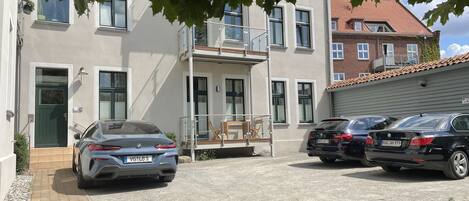 Ein Wohnhaus mit gepflasterter Auffahrt, zwei parkenden Autos und einem Balkon mit Gartenmöbeln.
