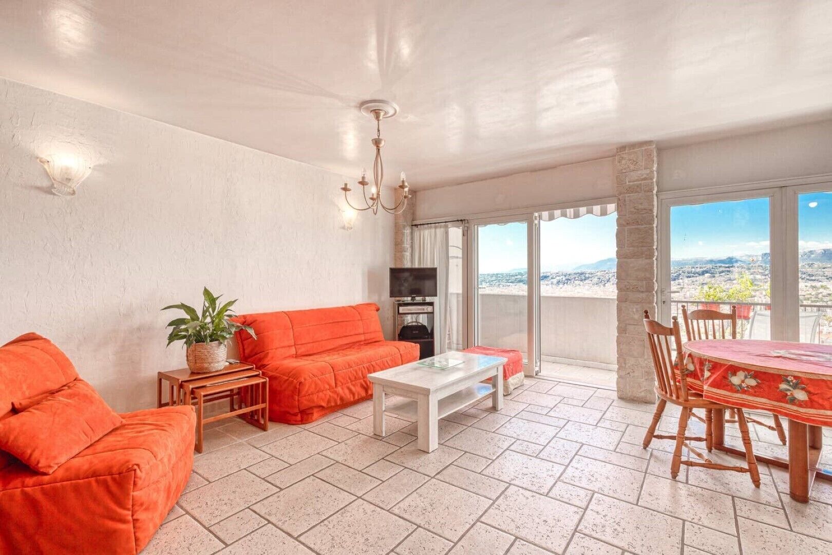 Un salon lumineux avec un canapé, une table basse et une table à manger avec des chaises. On y trouve également un téléviseur et une plante sur une table d’appoint. La pièce est dotée d’une grande fenêtre offrant une vue sur la ville.