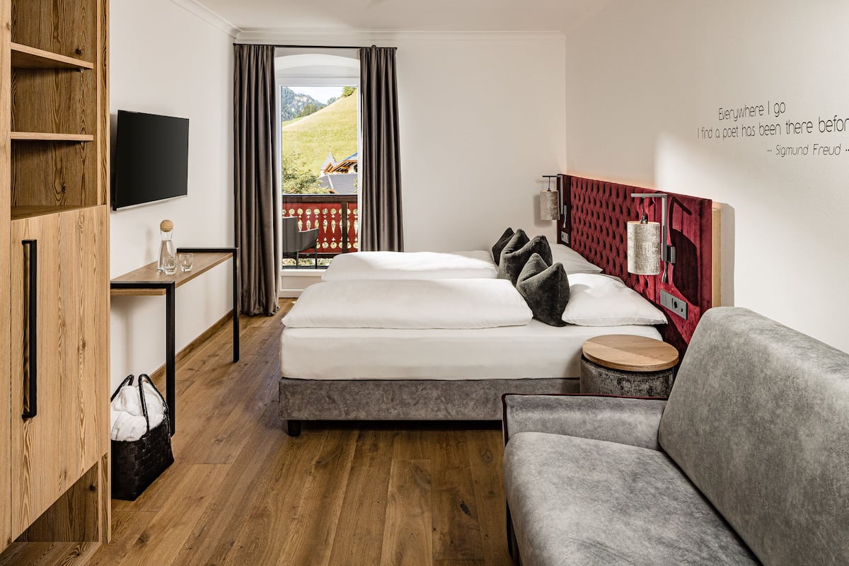 Et hotelværelse med en seng, en grå sofa, en træskab, et fjernsyn, et lille bord med et glas, samt udsigt til et bjerg og en rød bygning.