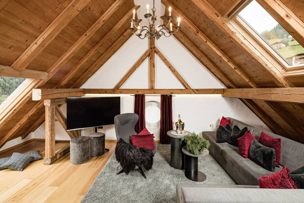 Et hyggeligt soveværelse på loftet med træbjælker, en lysekrone, et stort fladskærms-tv, en grå sofa med røde puder, en sort lænestol og et lille sofabord med en plante.