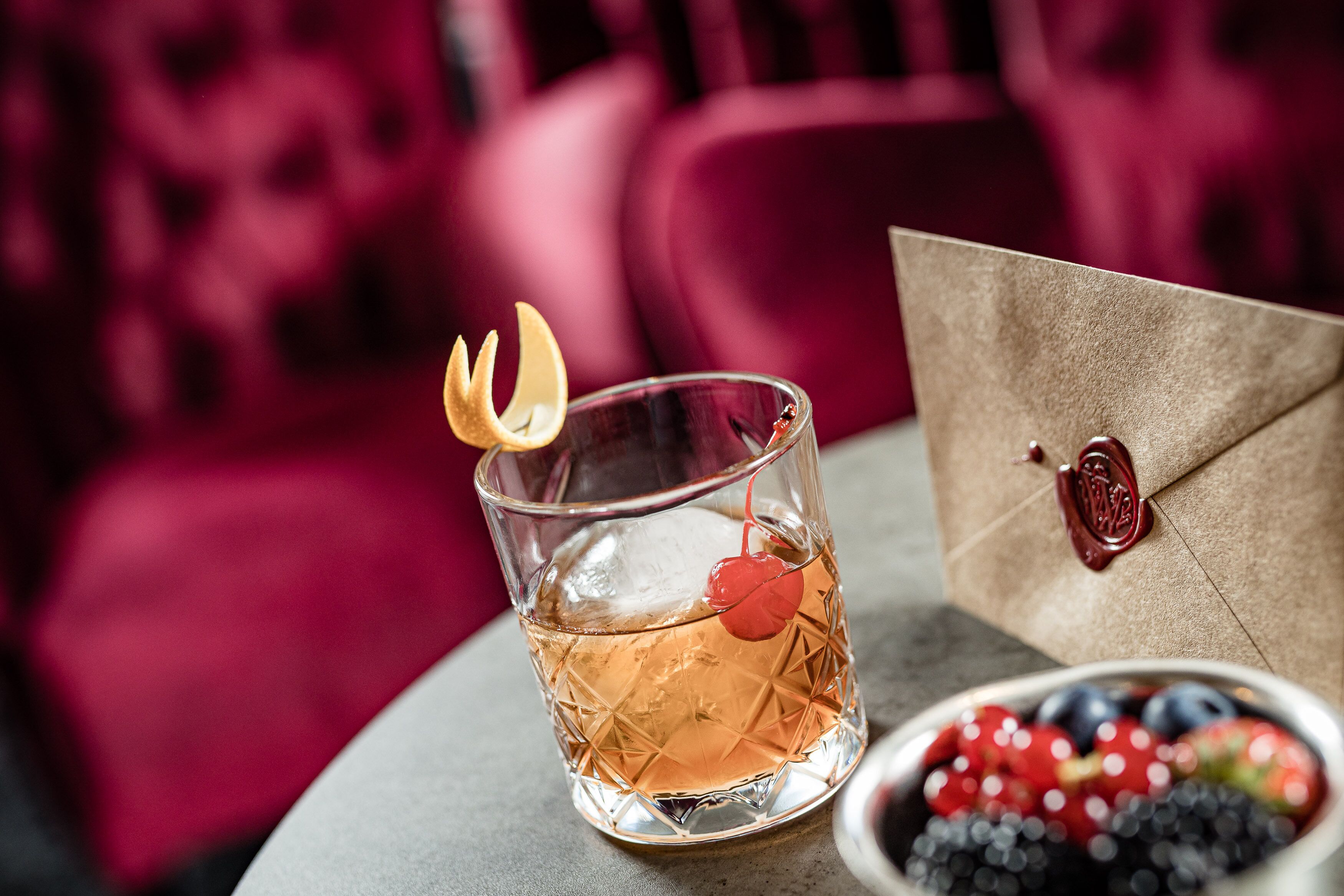 Un verre contenant un liquide ambré, garni d’une cerise et d’une tranche de citron, un bol de fruits rouges mélangés et une enveloppe brune scellée à la cire, le tout posé sur une table.