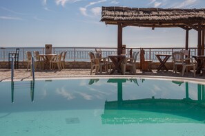 A pool with clear water, surrounded by chairs and tables, with a beach view in the background. - Korfiatis Apartments (Zakynthos)