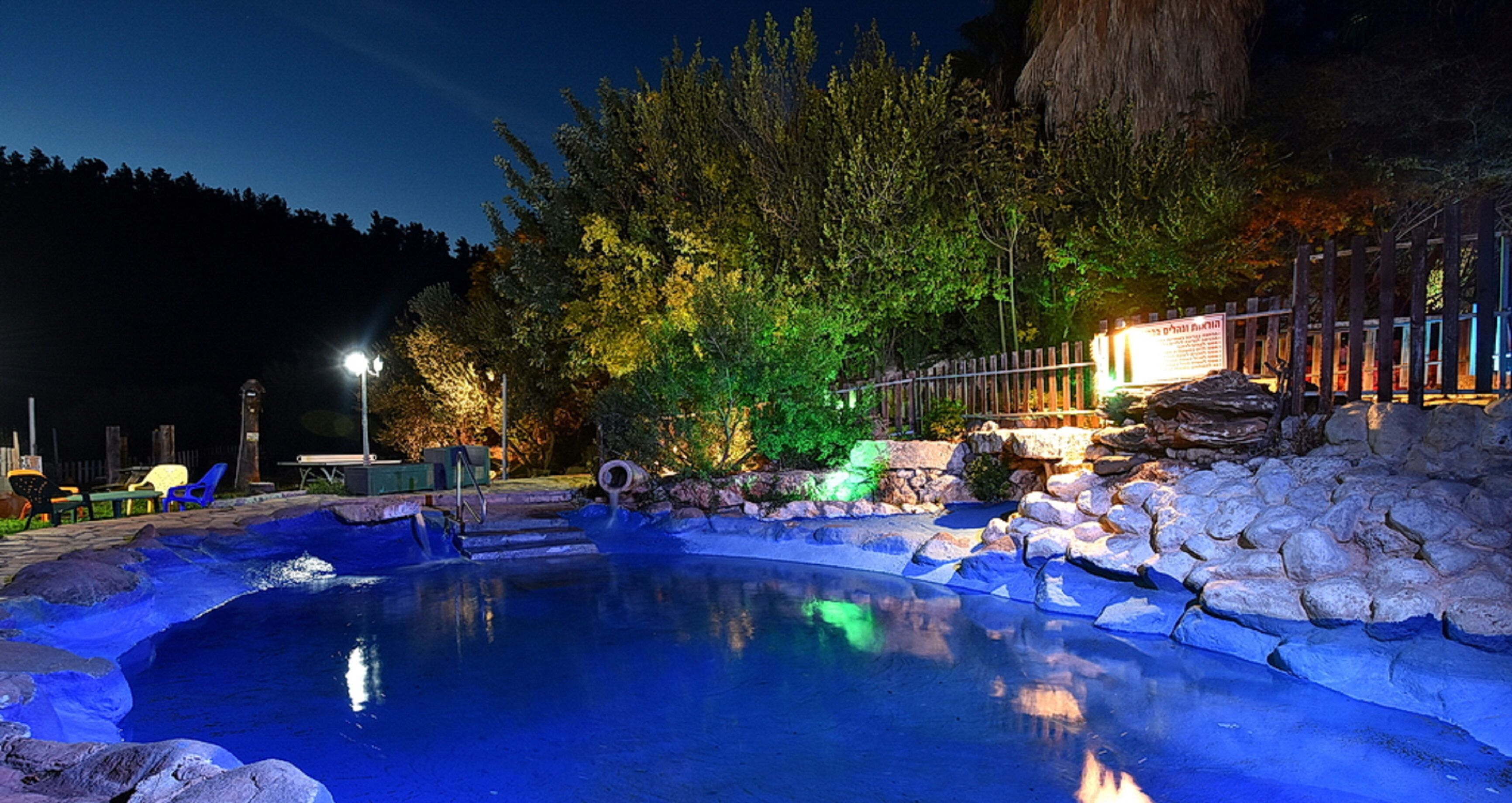 A nighttime outdoor pool area with a stone wall, a wooden fence, and trees illuminated by artificial lighting.