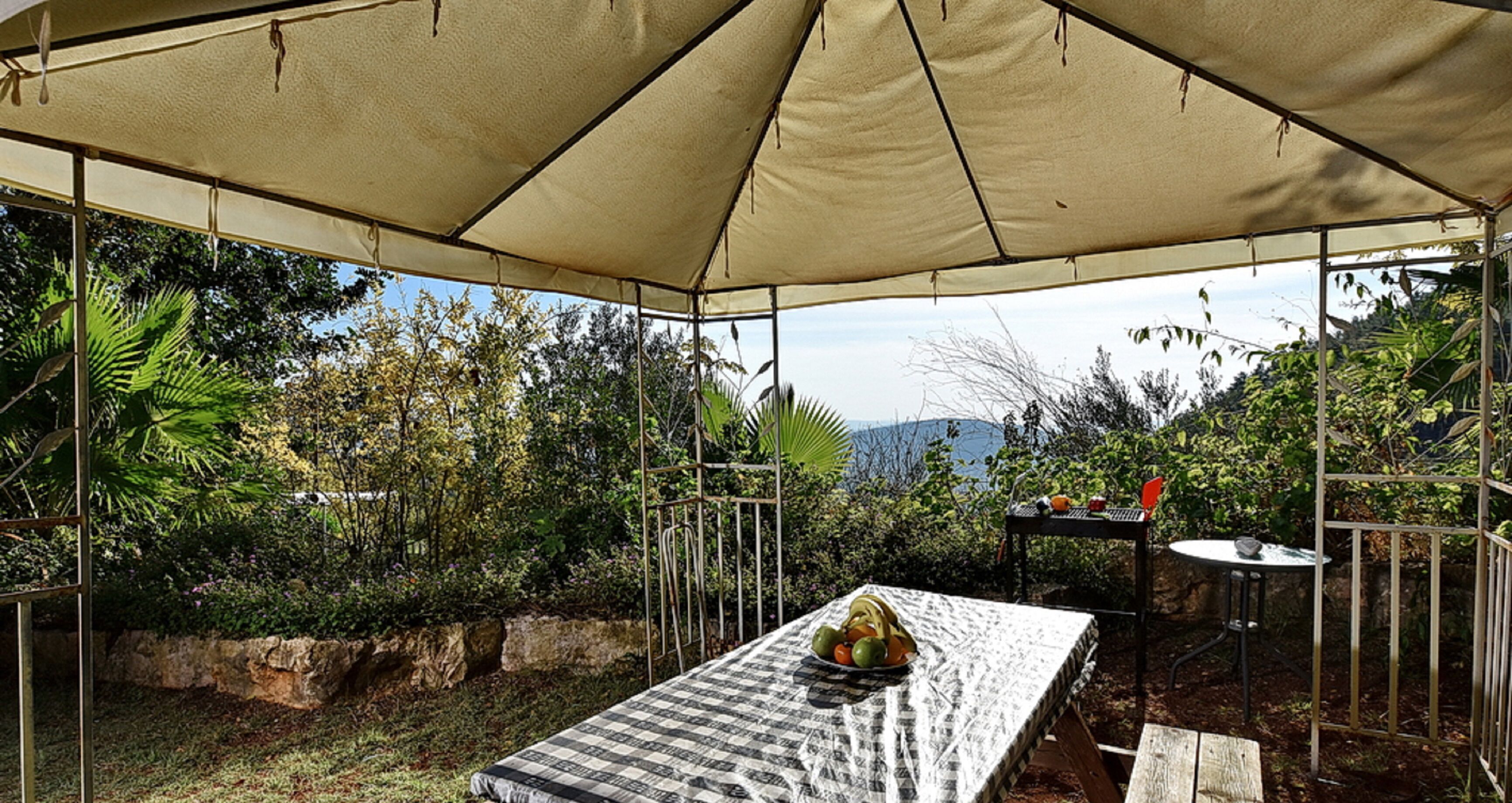 A covered outdoor seating area with a table set for two, surrounded by lush greenery and a view of distant hills.