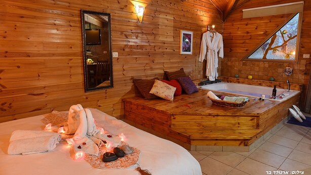 A wooden cabin interior with a large bed, a wooden bench, a bathtub, and a mirror. - Niflaot Hagalil (Merom HaGalil)