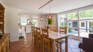 Un comedor amplio con sillas de madera y una mesa grande, una zona de cocina con armarios blancos y ventanas grandes que ofrecen vistas a la vegetación exterior.