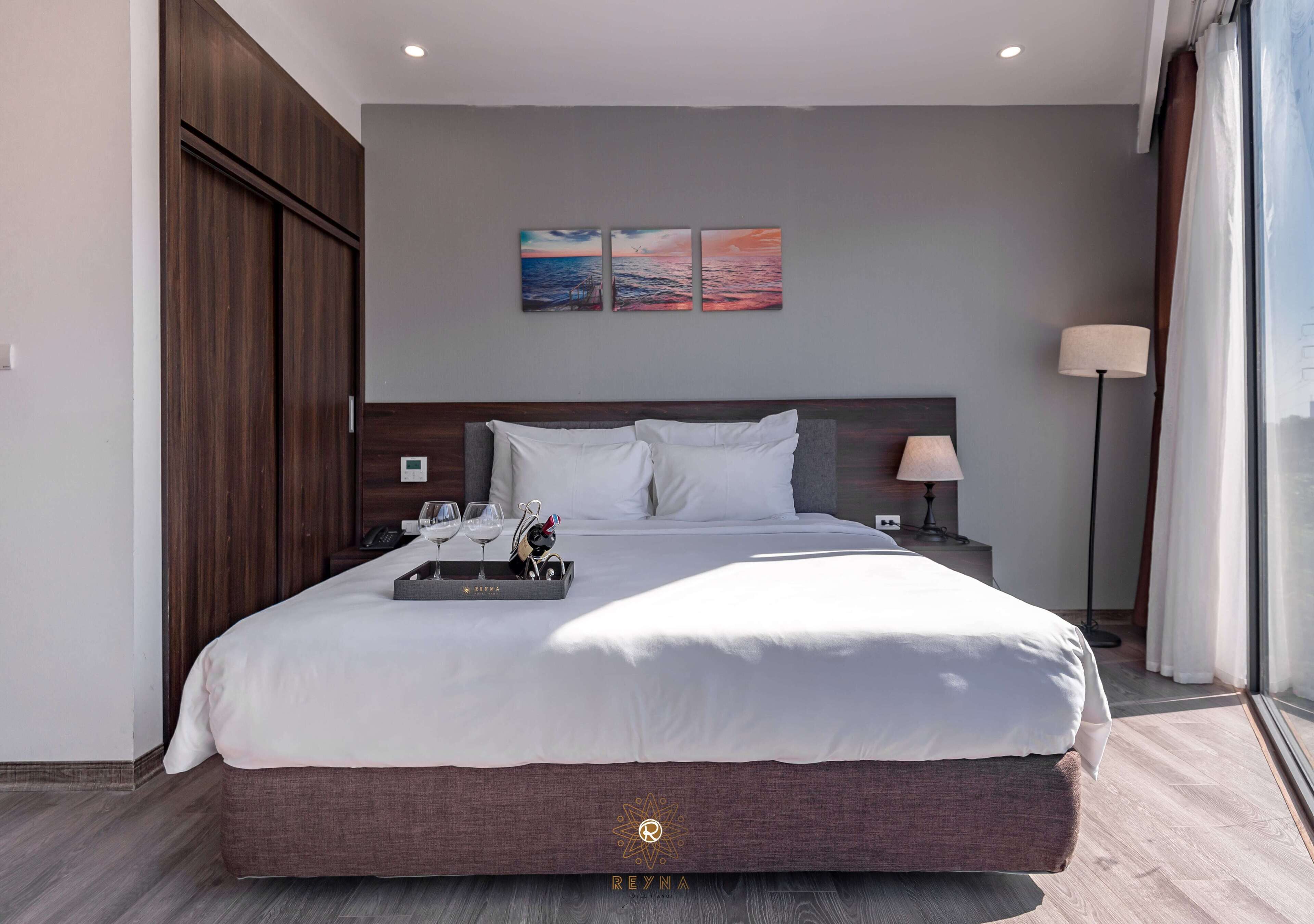 A modern hotel room with a large bed, a nightstand, a lamp, and a view of the sea through the window.