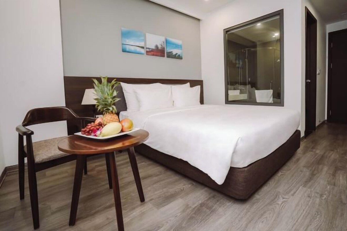 A hotel room with a bed, a small table with fruit, a chair, and a large window.