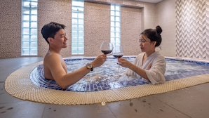 Um casal em uma banheira de hidromassagem com piso de azulejos, cercado por grandes janelas e uma parede com textura.