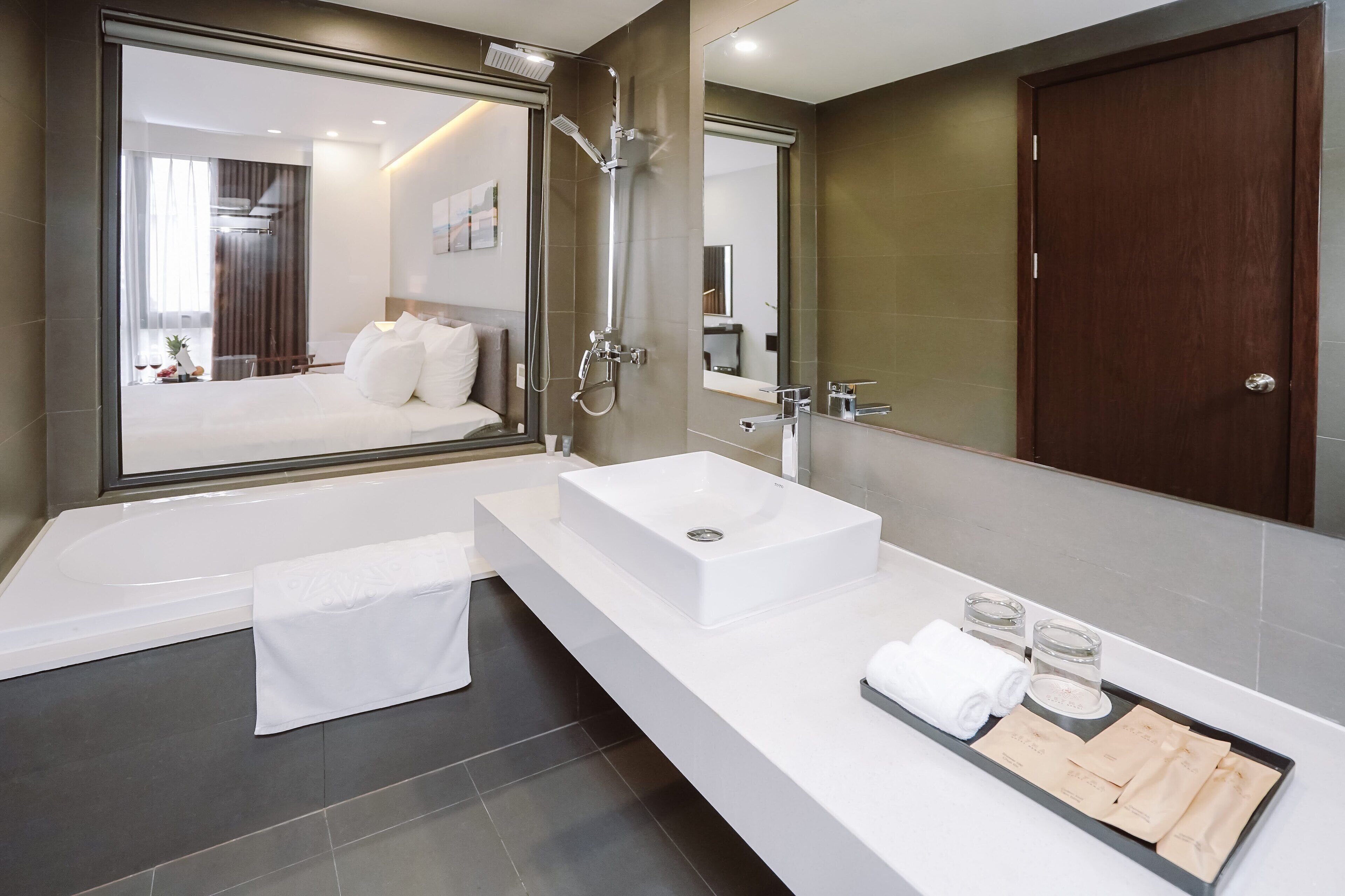 Un baño moderno con un espejo grande, una bañera, un lavamanos blanco y una bandeja con toallas y artículos de aseo.