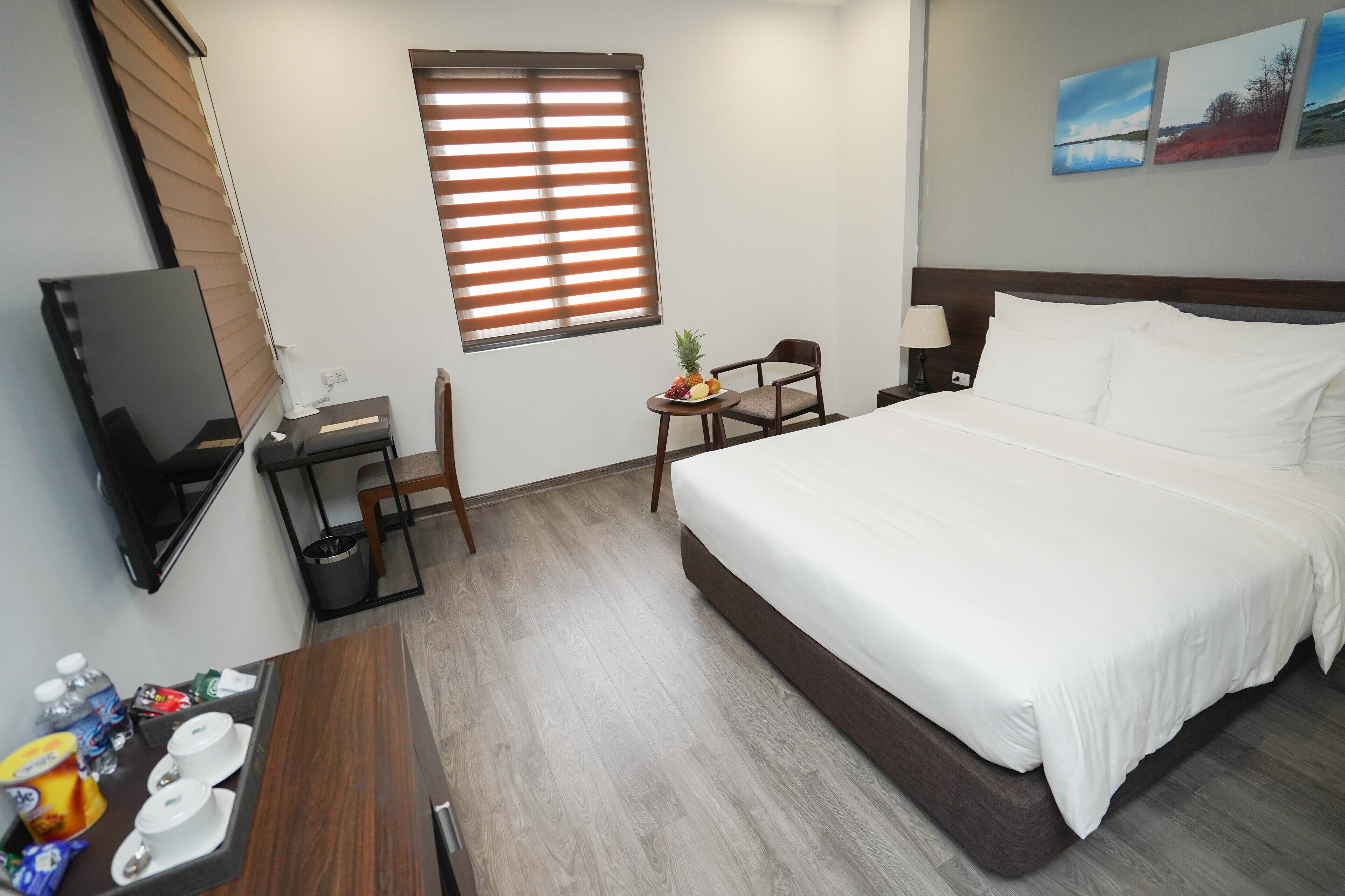 Habitación de hotel con una cama grande, televisión, una mesita con frutero, una silla y ventana con persianas.