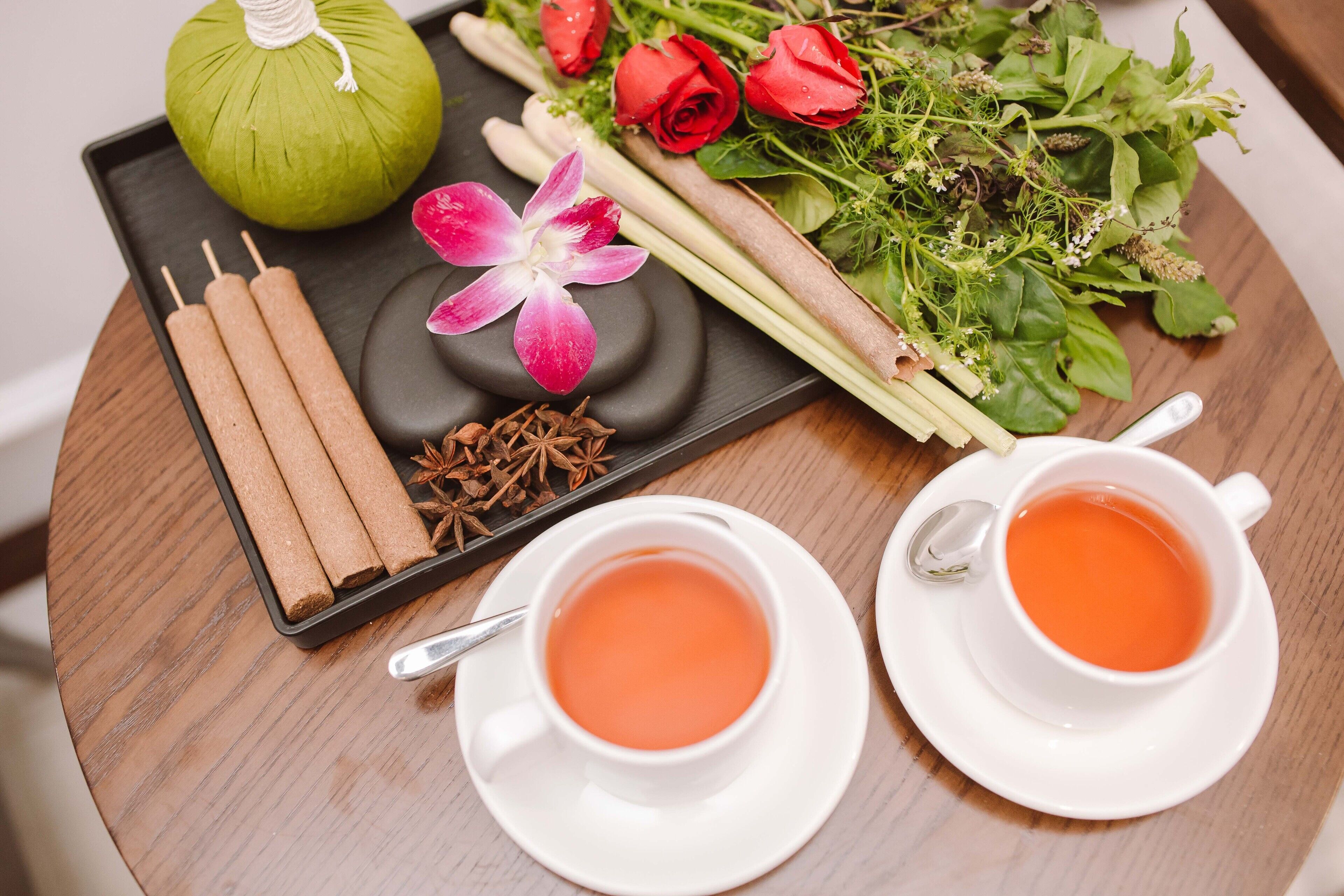 A tray with tea, a green fruit, rose petals, and a flower on a wooden table.