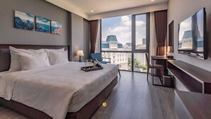 Quarto de hotel moderno com uma cama grande, uma escrivaninha e uma televisão. Há três quadros na parede e vista para a cidade pelas janelas.