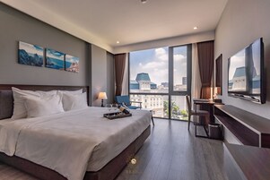 A modern hotel room with a large bed, a desk, and a TV. There are three paintings on the wall and a view of the city through the windows.