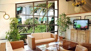 A living room with a sofa, coffee table, and wicker chairs. There is a television on a wooden cabinet, and a large window with a view of greenery and buildings. - Hotel Valle Verde (Marciana)