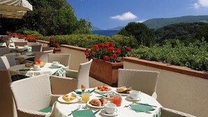 An outdoor dining area with wicker chairs and tables, a view of the sea, and red flowers in planters. - Hotel Valle Verde (Marciana)
