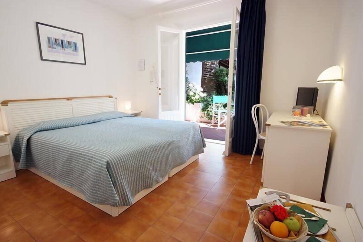 A bedroom with a bed, bedside table, and a desk with a lamp. There is a window with curtains, a door leading outside, and a small balcony with plants.