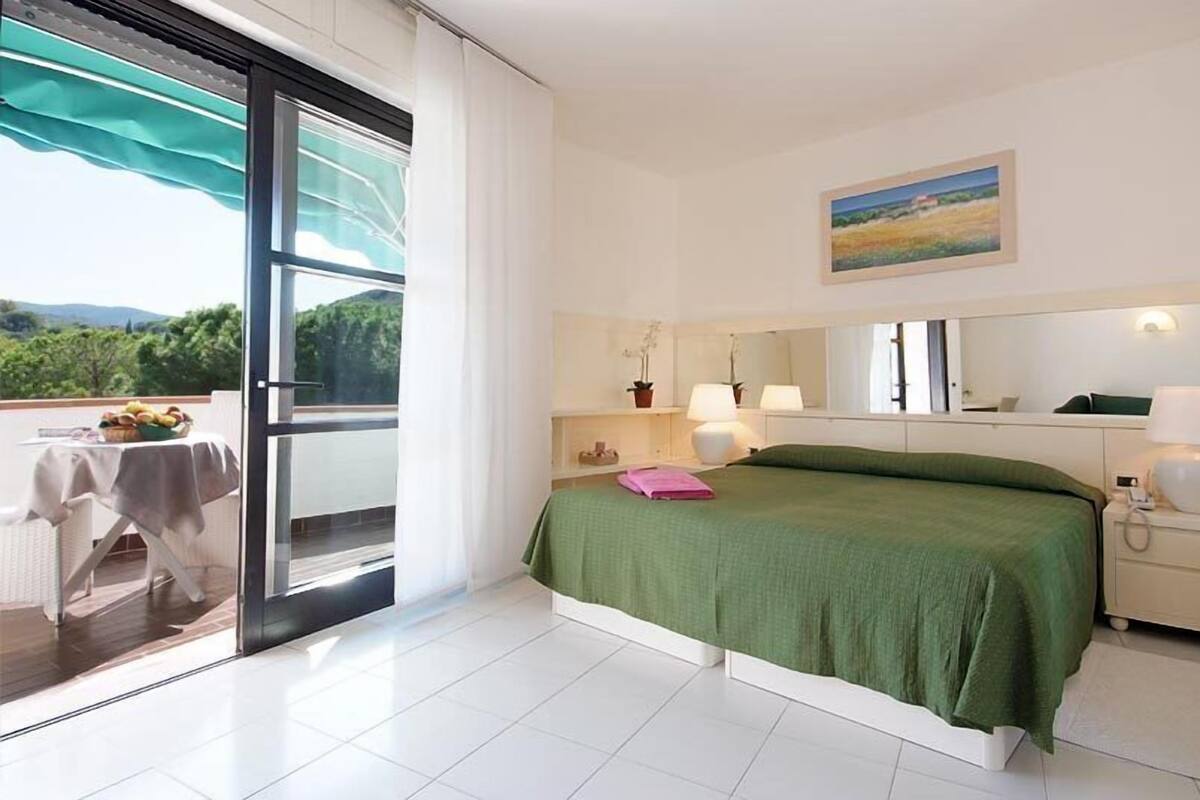 A hotel room with a balcony, a bed with a green bedspread, a bedside table with a lamp, a nightstand, and a painting on the wall.