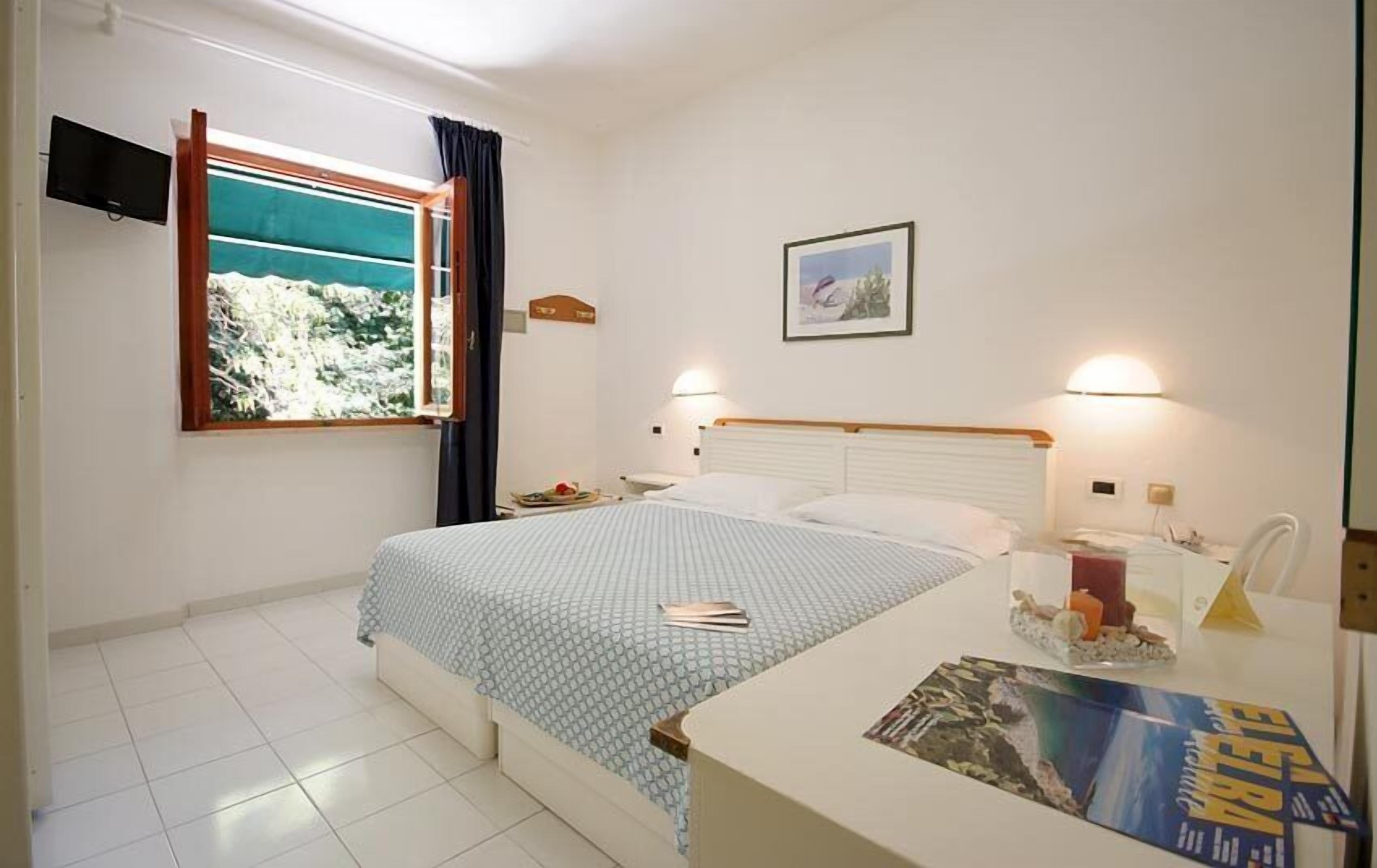 Un dormitorio con cama, una ventana con cortinas, una mesita de noche con lámpara y un cuadro en la pared.