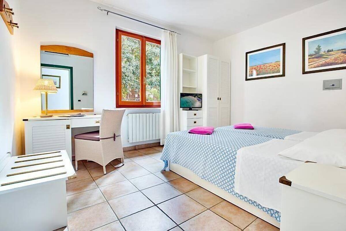 A bedroom with a bed, a desk, a chair, a radiator, a TV, and two paintings on the wall.