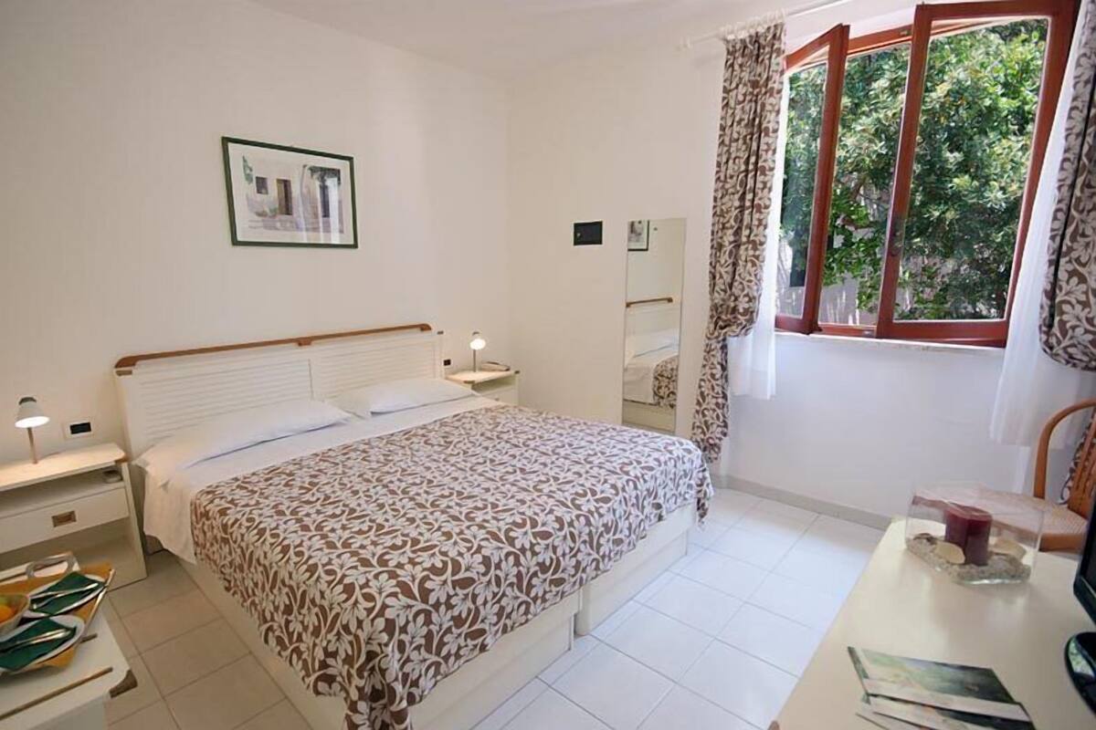 A bedroom with a bed, bedside tables, a desk, a chair, a window with curtains, and a framed picture on the wall.