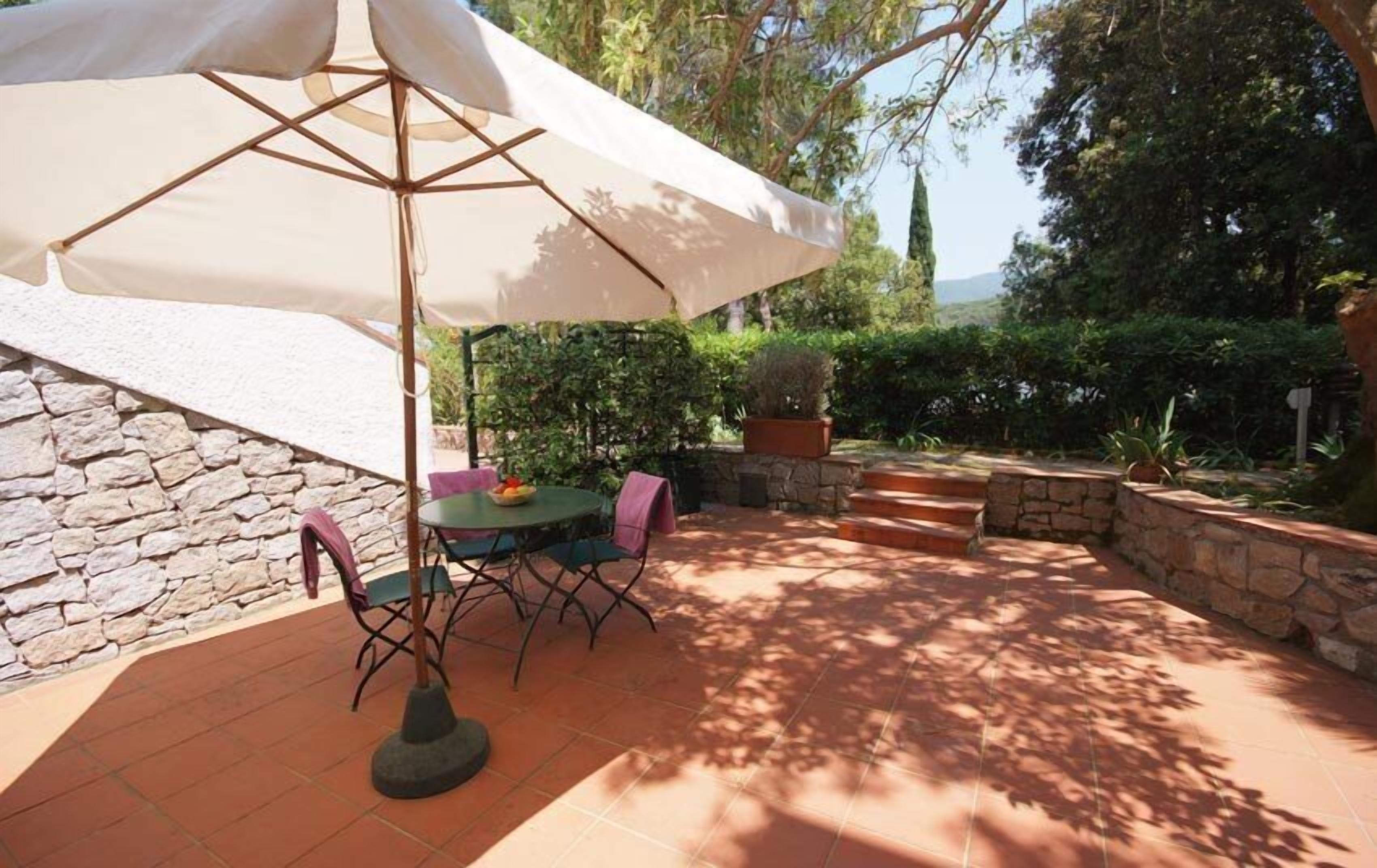 An outdoor patio with a table and chairs under a large umbrella, surrounded by stone walls and greenery.