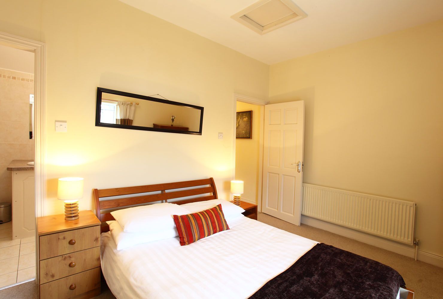 A bedroom with a wooden bed, two bedside tables with lamps, a mirror above the bed, a radiator, and a door to another room.