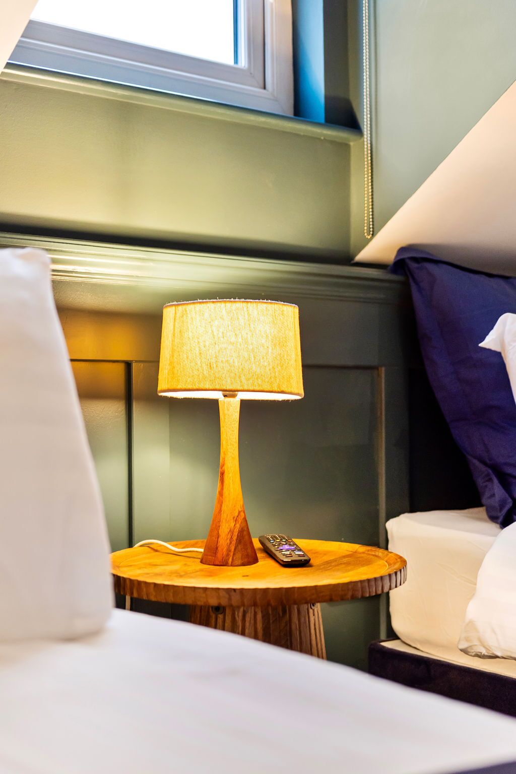 A wooden table with a lamp and a remote control, a bed with white bedding, and a window with blinds.