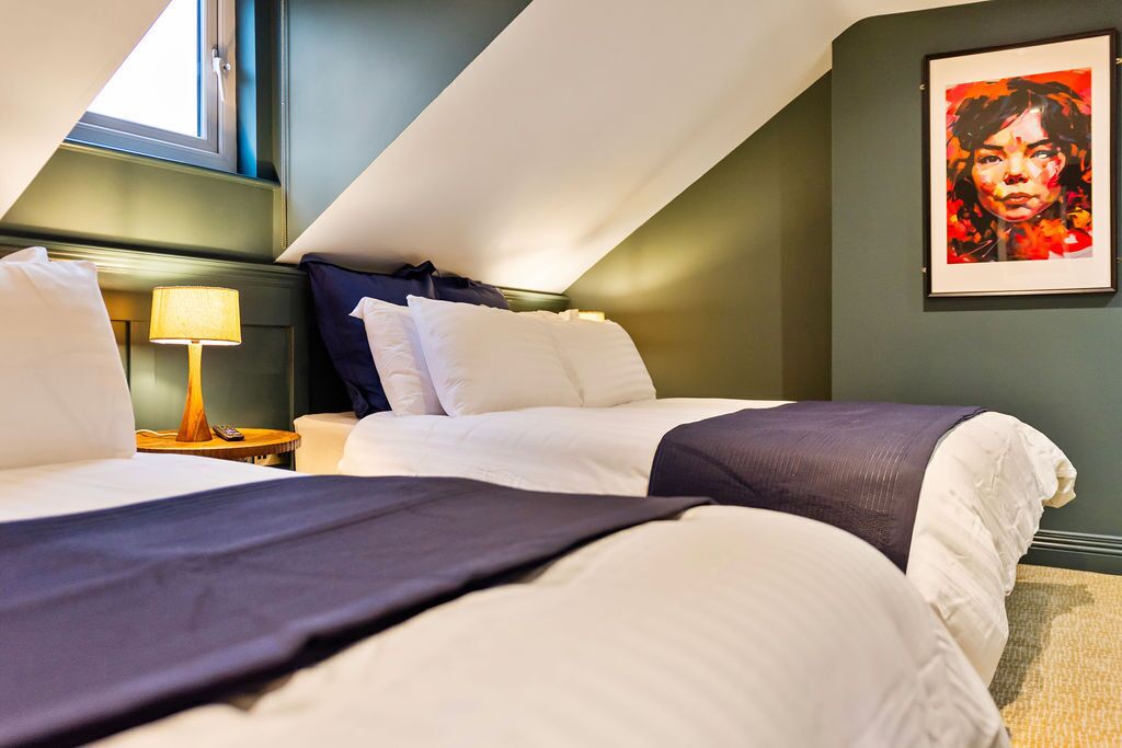 A hotel room with two beds, a nightstand, a lamp, and a framed artwork on the wall.
