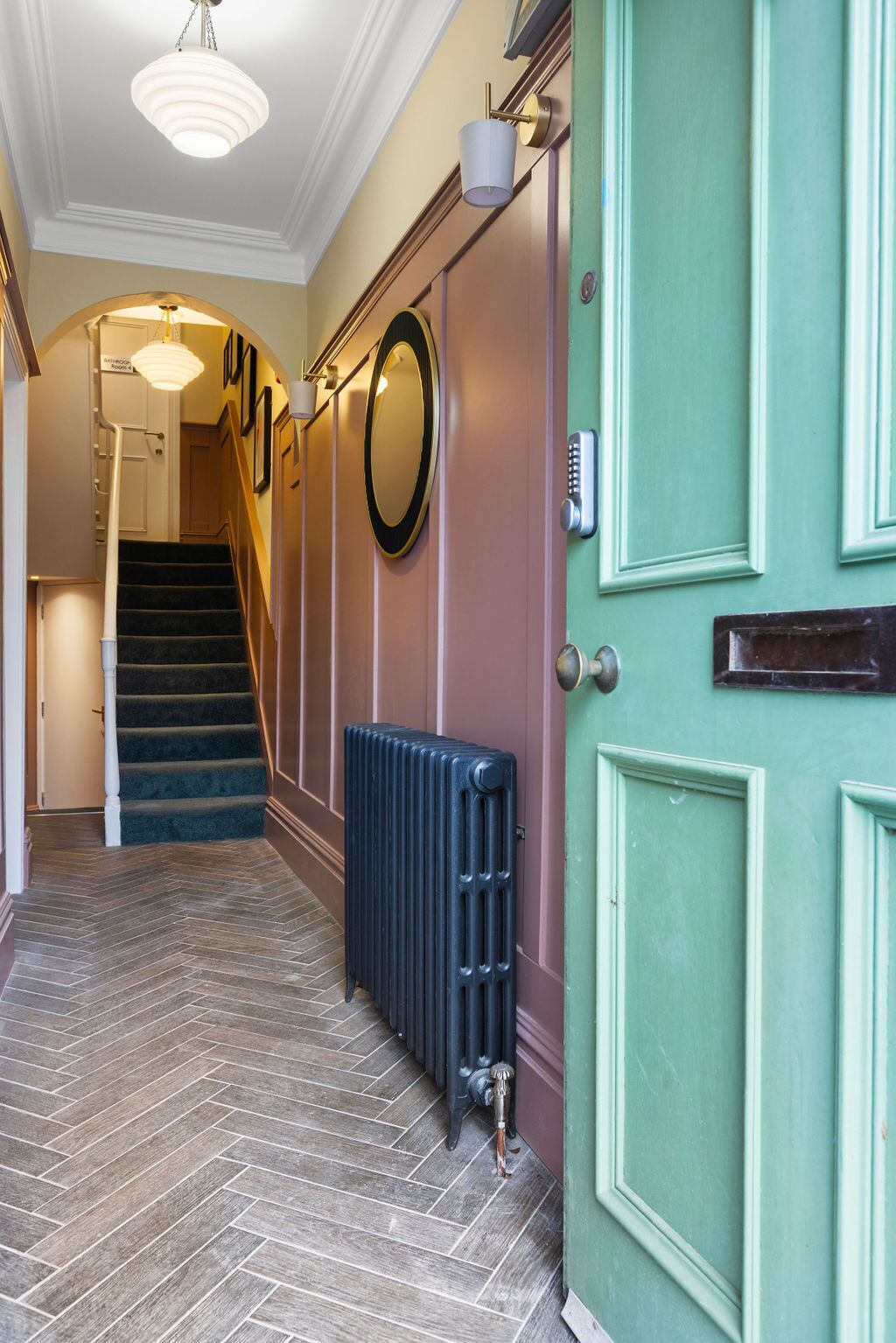 Um corredor com piso em padrão herringbone, um radiador, uma porta verde e uma escadaria que leva a outro cômodo.