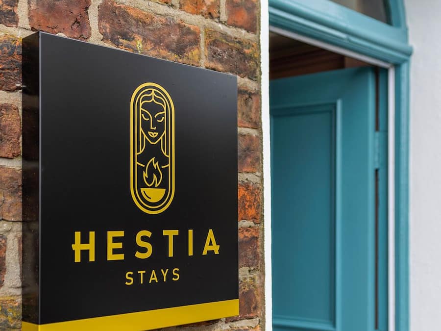 Uma placa preta com um logotipo amarelo e a inscrição 'HESTIA STAYS' em uma parede de tijolos.