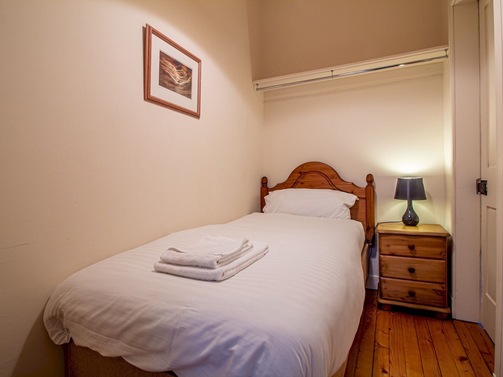 Una cama bien tendida con sábanas blancas y una toalla doblada, una mesita de noche de madera con una lámpara, y un cuadro en la pared.