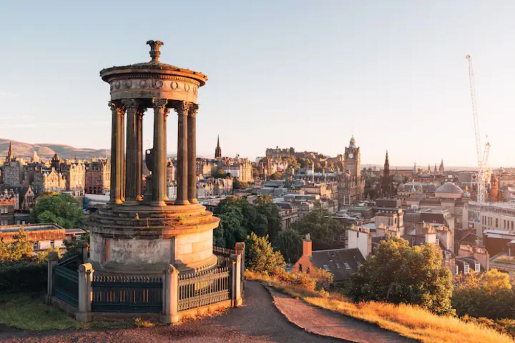 A historical monument with columns, surrounded by a pathway and greenery, overlooking a cityscape with various buildings and a clock tower.