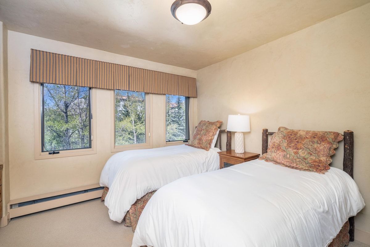 A bedroom with two beds, a window with a view of trees, and a bedside table with a lamp.