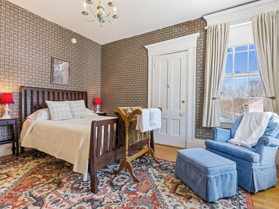 Un dormitorio con cama, una silla, una mesita, un tapete estampado y una ventana con cortinas.