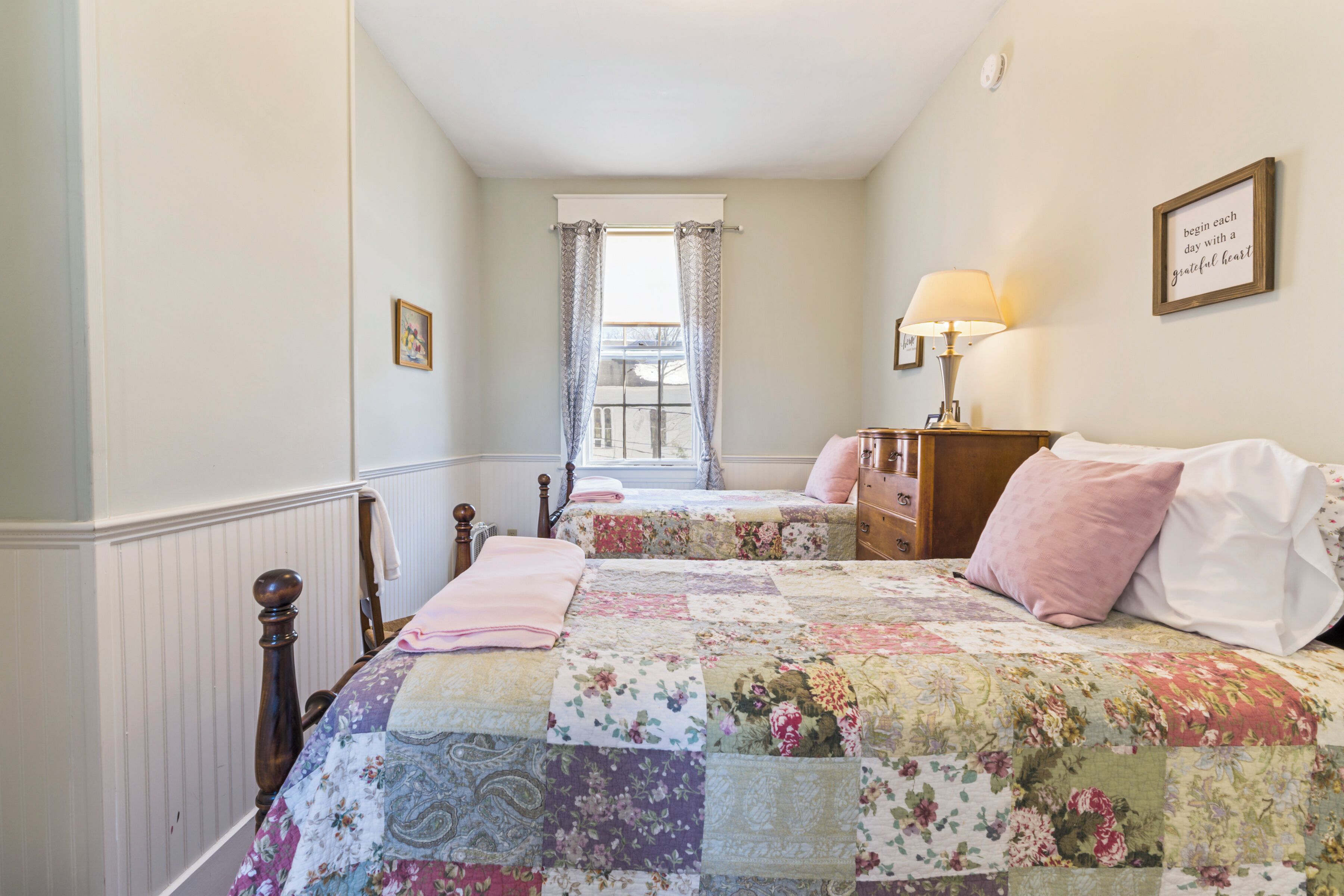 A bedroom with two beds, a window with curtains, a bedside table with a lamp, and a framed quote on the wall.