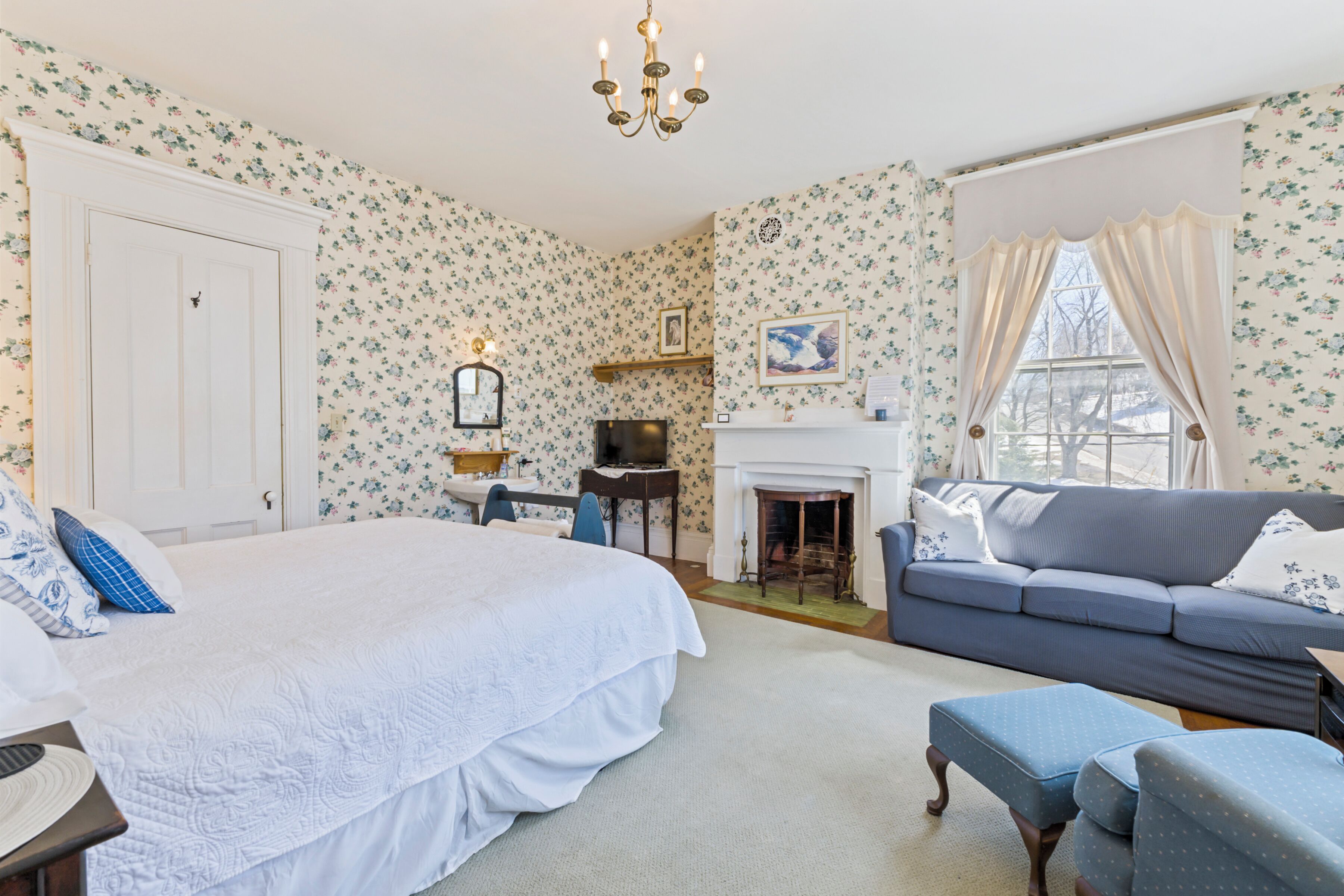 A bedroom with a large bed, a sofa, a fireplace, and floral wallpaper.