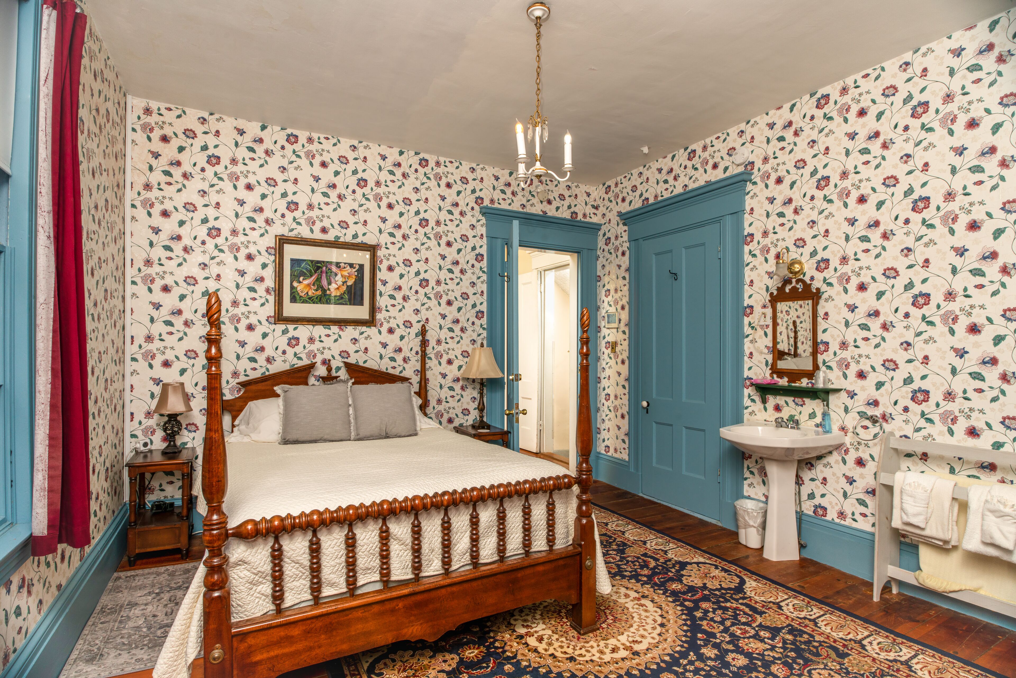 Une chambre à coucher avec un lit à baldaquin en bois, un papier peint à motifs floraux, un lustre, une table de chevet avec une lampe et une commode.