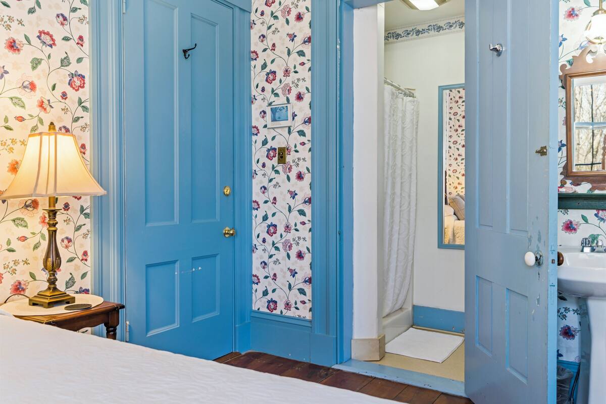 A room with a blue door, floral wallpaper, a bedside lamp, and a bathroom with a shower curtain.