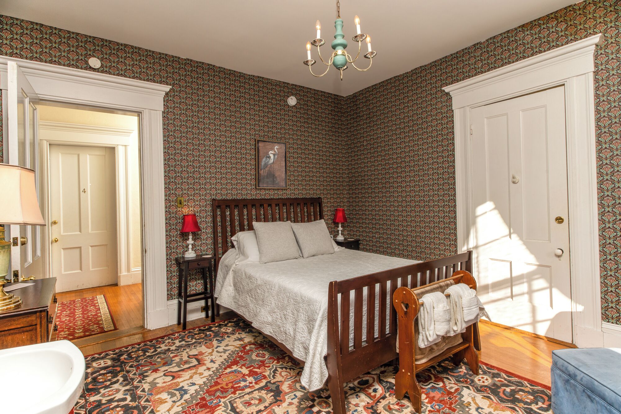 A bedroom with a four-poster bed, a chandelier, patterned wallpaper, and a wooden bench with towels.