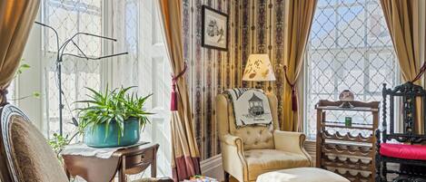 A room with a patterned wallpaper, a chair, a table with a lamp, a potted plant, and a window with curtains.